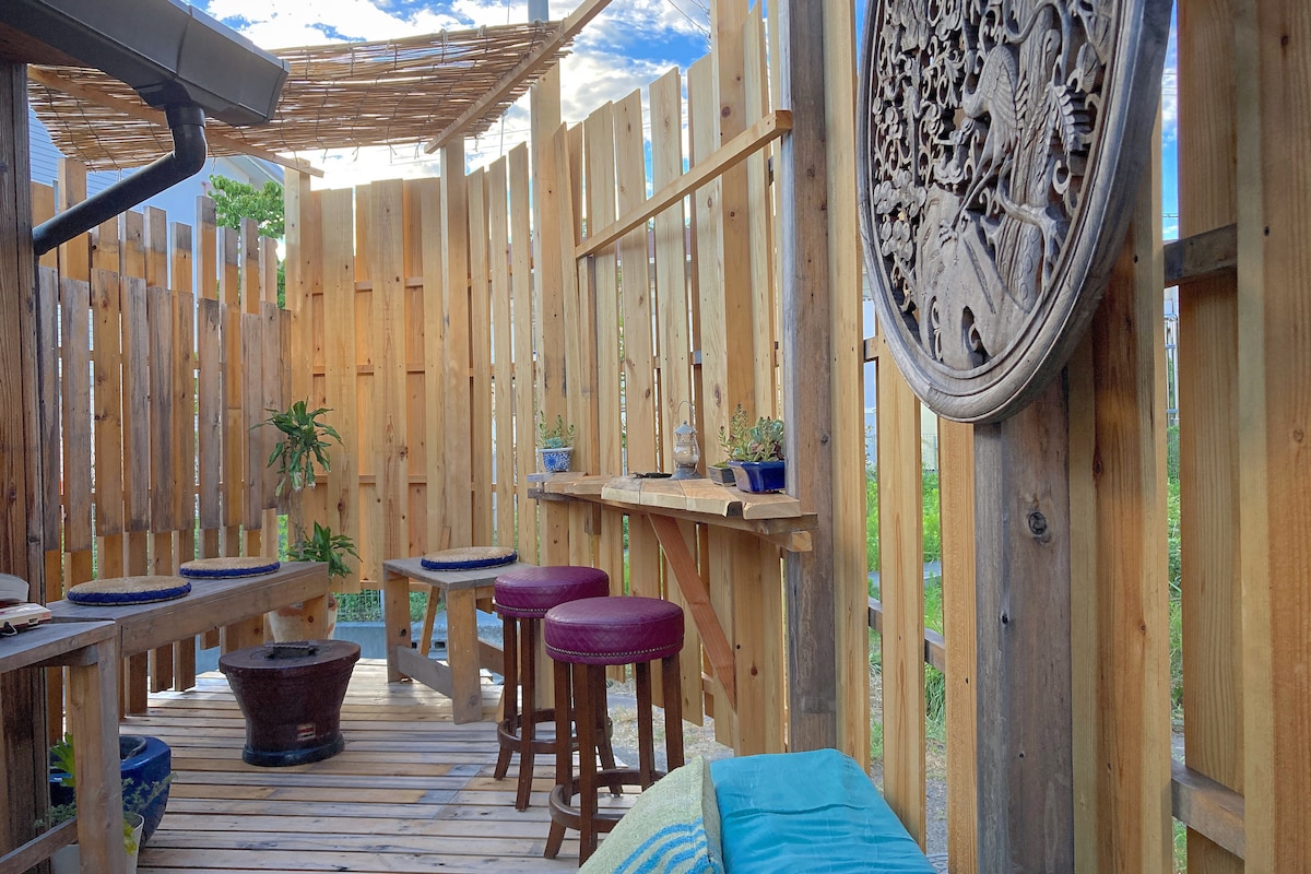 An outdoor wooden deck area is surrounded by slatted fencing, creating a private and cozy atmosphere. Wooden seating includes two tall stools and several low tables. Planters with greenery add a touch of nature, while decorative elements are integrated into the woodwork.