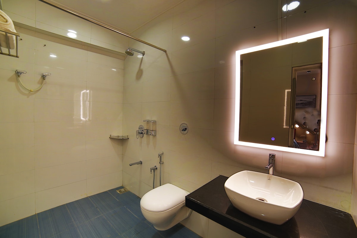 A modern bathroom features sleek white tiles and a contemporary design. An illuminated mirror is mounted above a minimalist sink on a dark countertop. A toilet is positioned beside a walk-in shower, which includes a glass partition and overhead shower fixture.