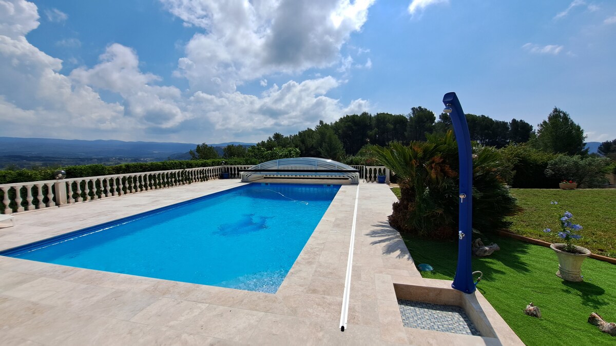 A large, secured swimming pool is set against a backdrop of vibrant greenery and distant hills. The pool area is surrounded by elegant stone tiles, with lounge chairs positioned nearby. Towering trees and a colorful flower pot contribute to the serene outdoor environment.