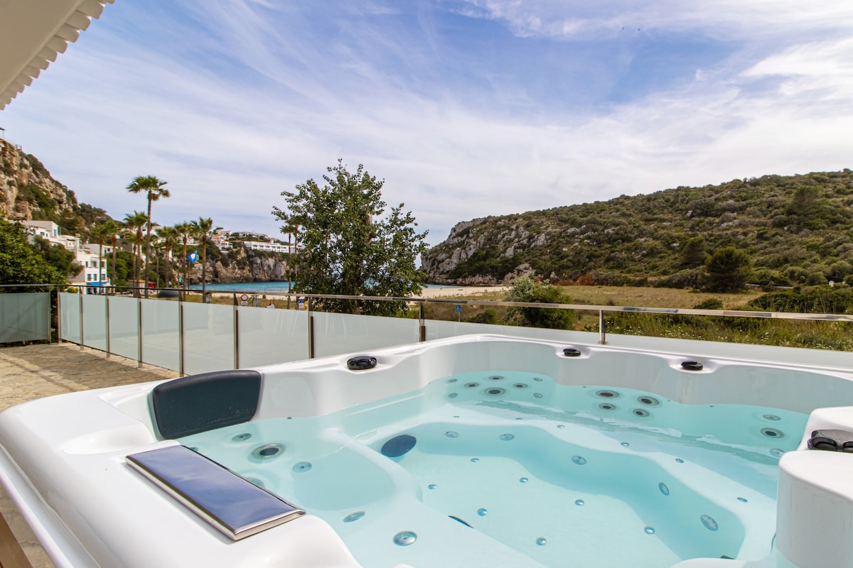 A spacious hot tub is situated on a terrace, overlooking a serene landscape of hills and greenery. Tall palm trees and a clear sky can be seen in the background, providing a tranquil outdoor setting.