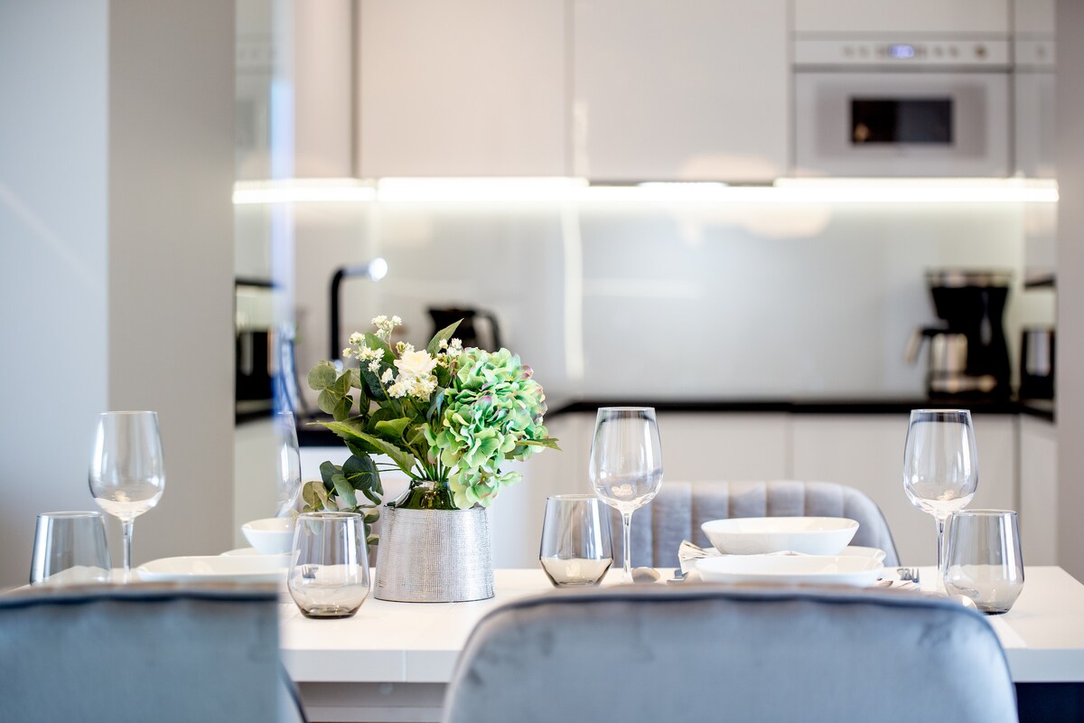 A modern dining area is presented, featuring a sleek white table set with elegant glassware and dishes. A small vase with fresh greenery adds a touch of nature. The backdrop displays a contemporary kitchenette, equipped with appliances and clean lines.