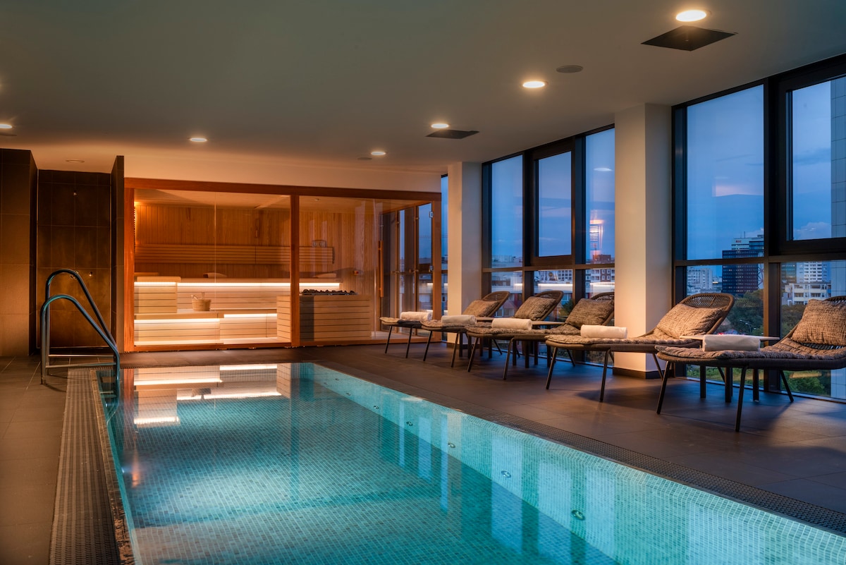 An indoor swimming pool area features a sleek design with a mosaic-tiled pool reflecting soft lighting. Lounge chairs are positioned along the edge, offering a tranquil space for relaxation. Panoramic windows provide views of the outside, enhancing the serene atmosphere.