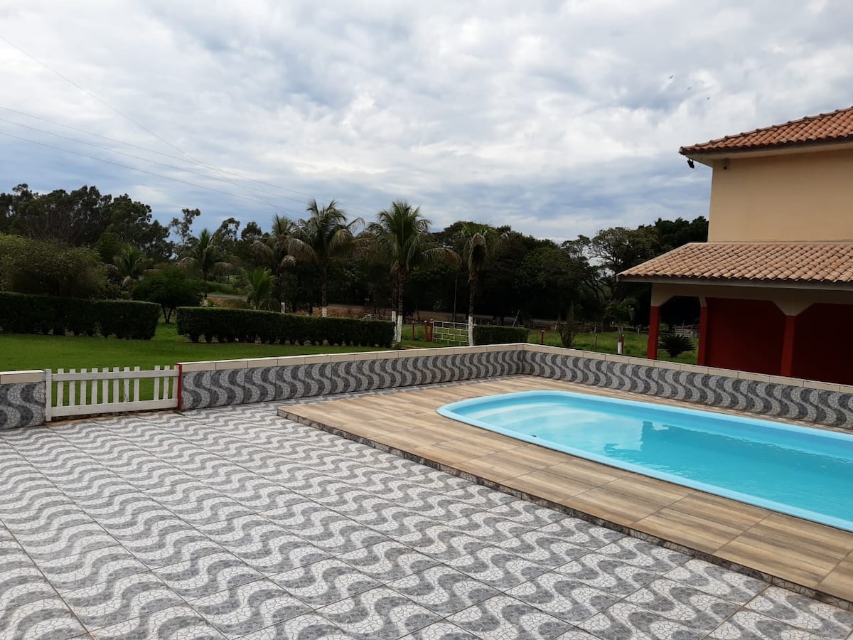 An outdoor area features a sparkling swimming pool surrounded by a patterned tile deck. Lush greenery and palm trees create a tranquil atmosphere, while a red structure with a tiled roof is visible in the background.