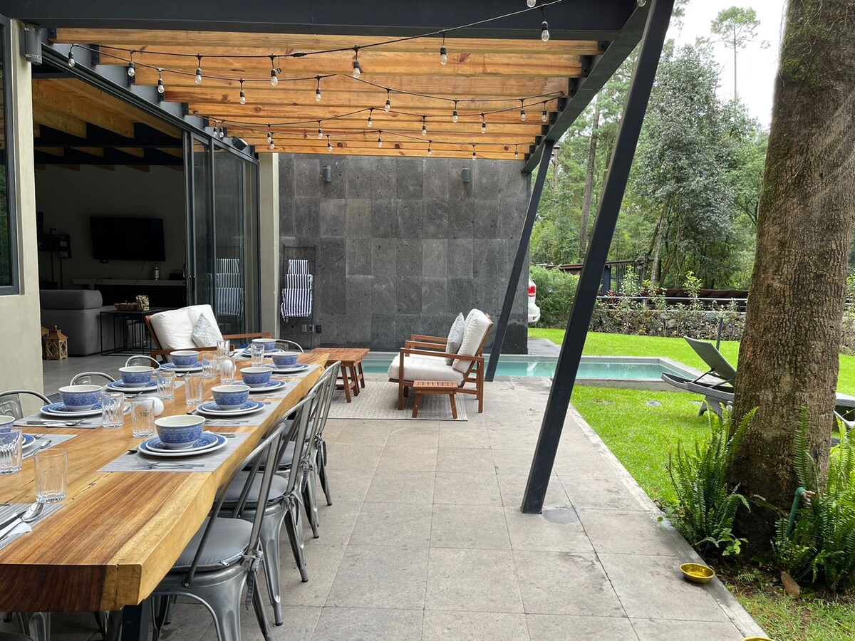 A spacious terrace is shown, featuring a long wooden dining table set for twelve, accompanied by metal chairs. A cozy seating area includes a wooden sofa and armchairs. Soft string lights hang overhead, and a clear swimming pool is visible in the background, surrounded by greenery.