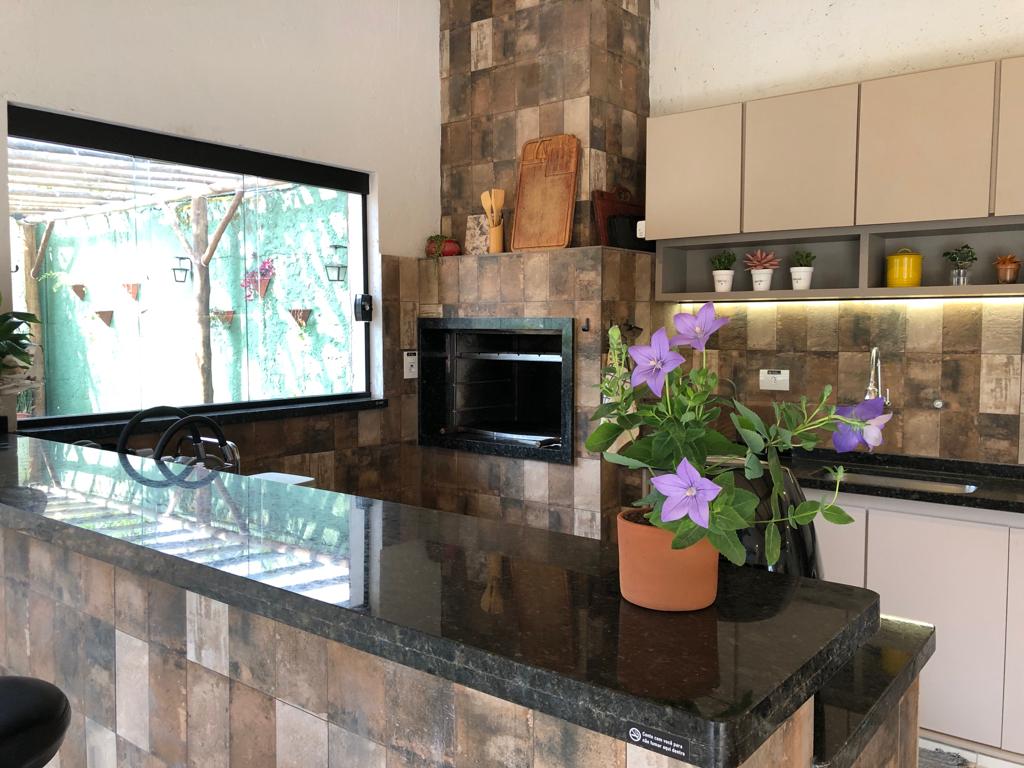 The kitchen features a dark granite countertop with a potted plant displaying purple flowers. A built-in oven is visible against a backdrop of textured stone tiles. Natural light enters through a window, revealing greenery outside.