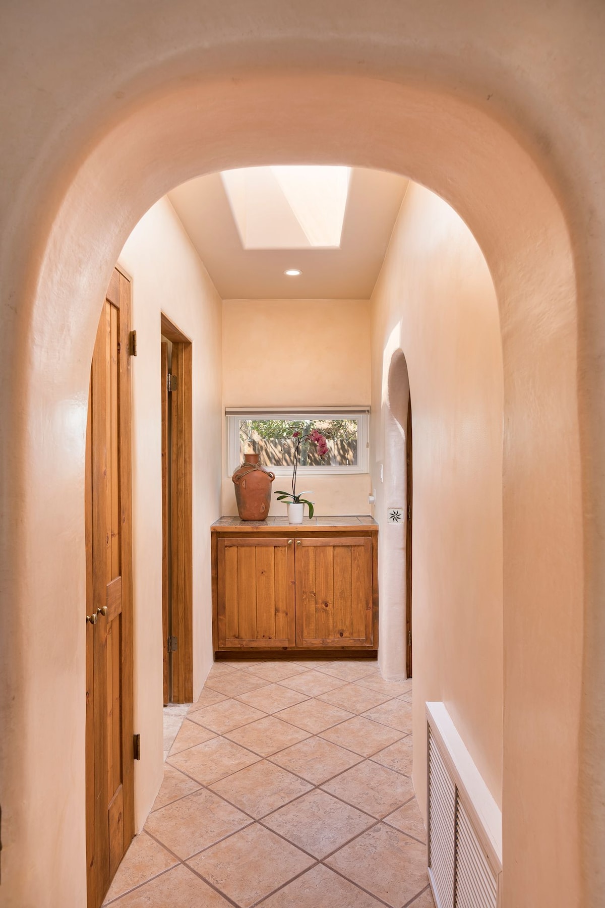 A light-filled hallway is framed by arched doorways and features warm, neutral walls. Natural light filters through a skylight, illuminating the tile flooring. A wooden cabinet sits against one wall, complementing the earthy tones throughout the corridor.