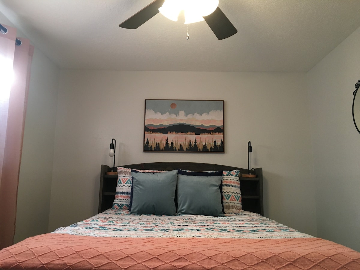 A cozy bedroom features a spacious bed adorned with patterned bedding and multiple decorative pillows. Soft lighting from two bedside lamps complements the artwork above the bed, which depicts a serene landscape. A ceiling fan adds to the room's comfort.