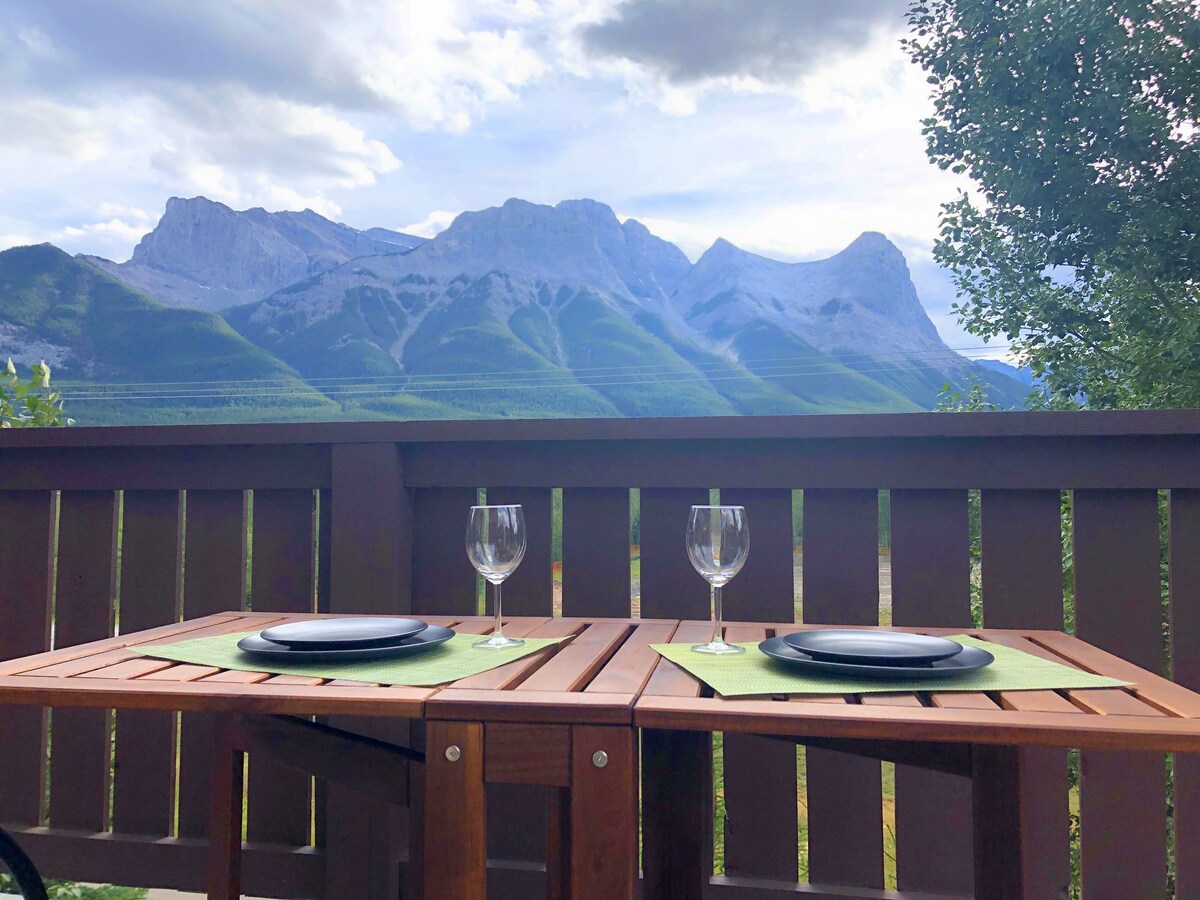 A private balcony table with green placemats is set for two, featuring empty glasses and plates. The backdrop boasts expansive mountain views under a cloudy sky, highlighting the natural beauty of the surroundings.