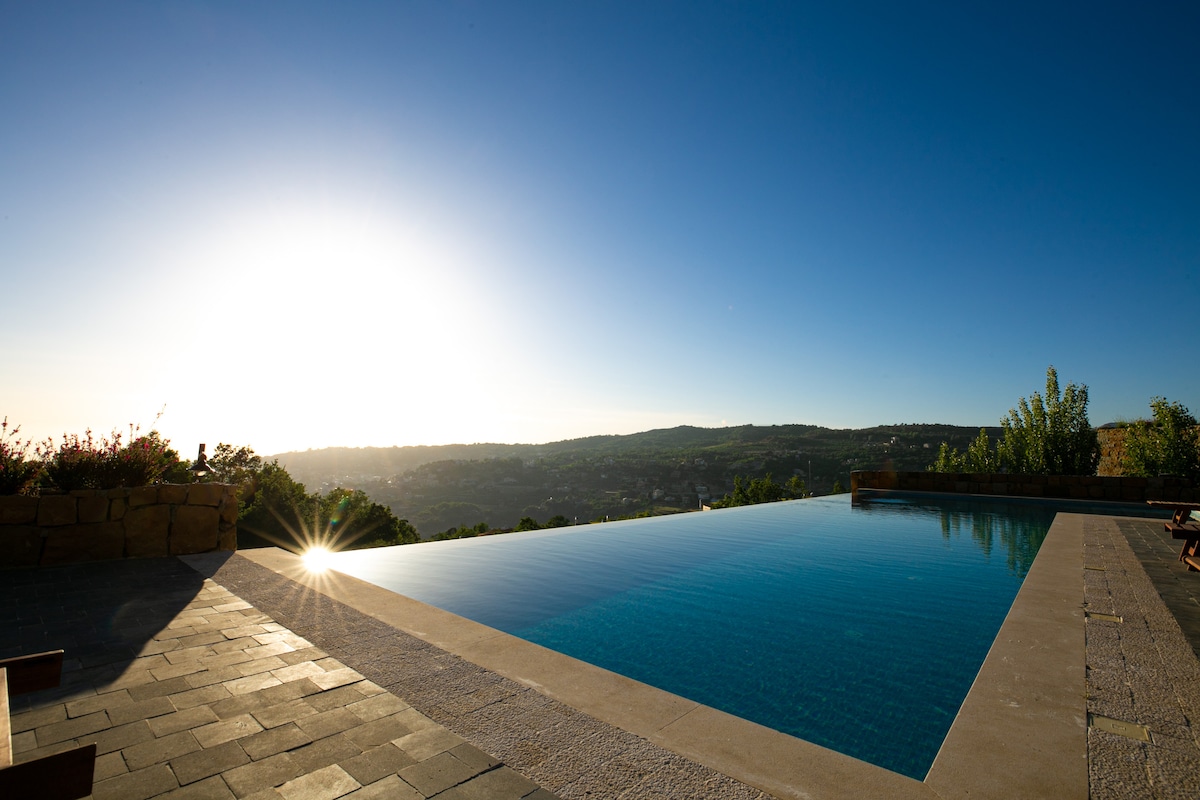 A serene infinity pool stretches into the horizon, reflecting the sunlight under a clear sky. Surrounding greenery and mountains create a picturesque backdrop, while the pool's smooth surface invites relaxation. The deck features simple lounge chairs positioned for comfortable seating.