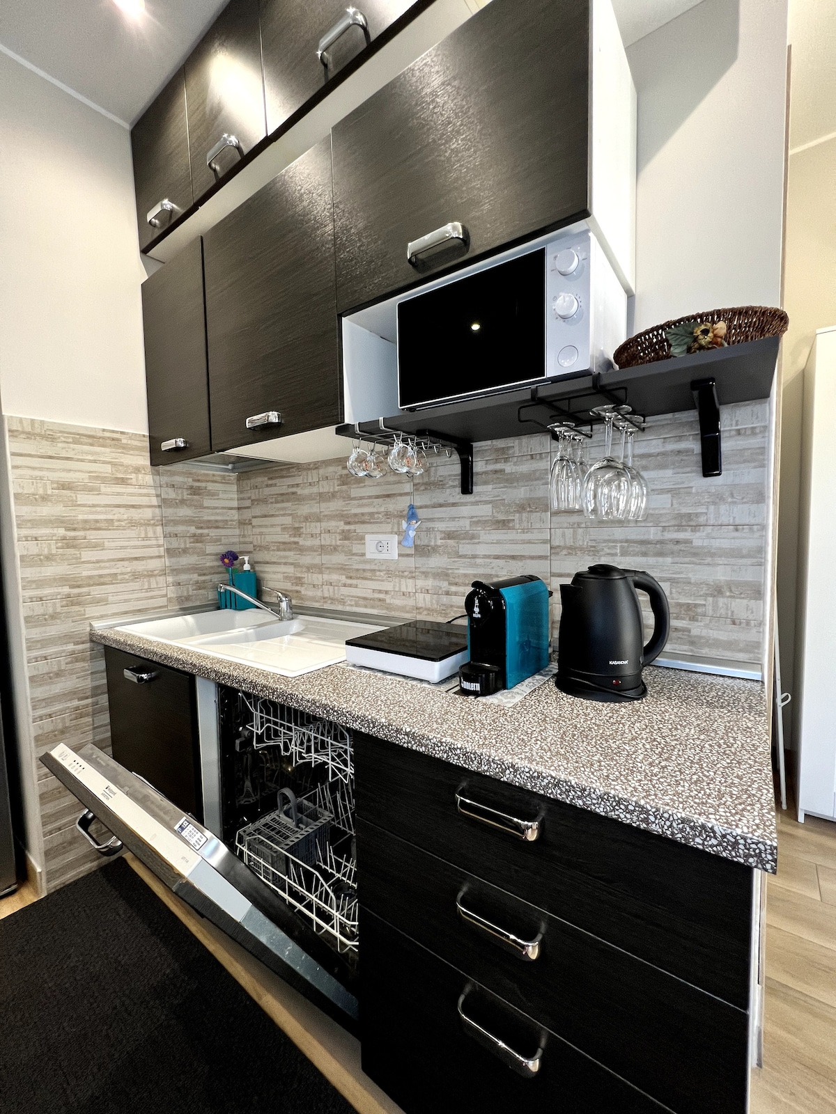 A modern kitchen space is equipped with sleek cabinetry and essential appliances. A dishwasher is conveniently installed beneath the counter, while a coffee maker, kettle, and microwave are placed on the counter. Glassware hangs above for easy access, and a sink is integrated into the design.
