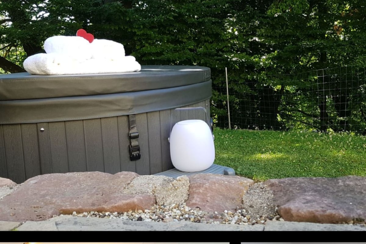 A whirlpool is surrounded by a stone wall, with fluffy towels resting on its edge. A small, illuminated speaker sits nearby, enhancing the outdoor relaxation area, which is framed by lush greenery.
