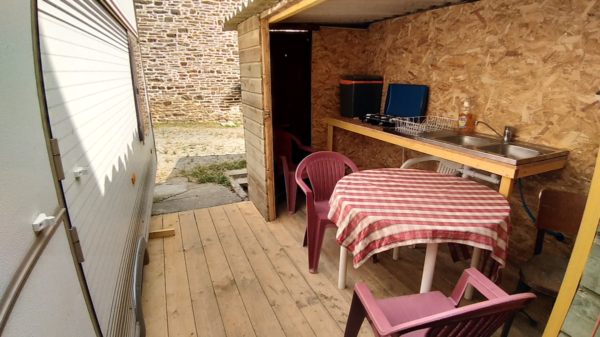 A simple outdoor area features wooden flooring with a checkered tablecloth covering a round table, accompanied by several plastic chairs. A compact kitchen area is visible with sinks and utensils. Surrounding walls have a natural wood finish, providing a rustic backdrop.
