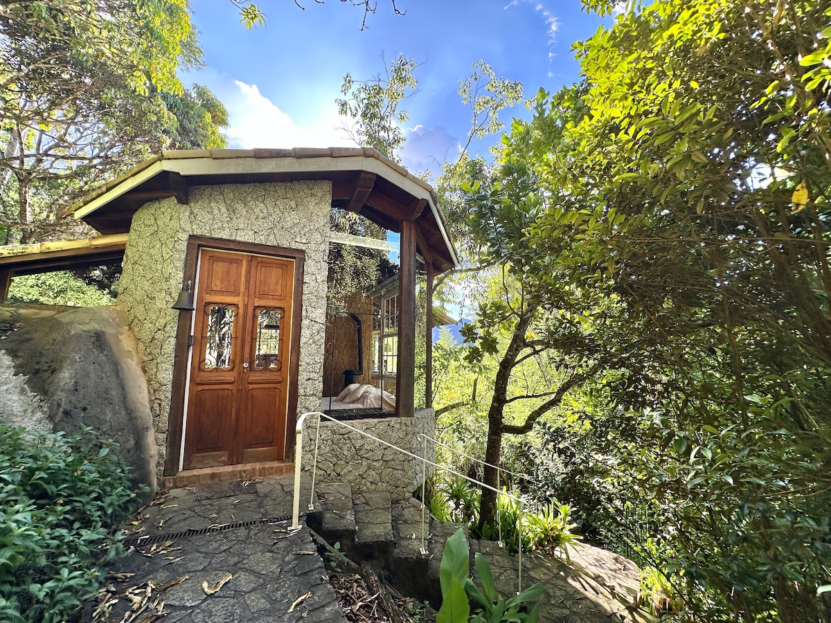 A charming stone chalet is set amidst lush greenery, with a wooden front door inviting entry. The large glass windows allow for natural light and views of the surrounding nature, enhancing the connection to the tranquil outdoor environment.