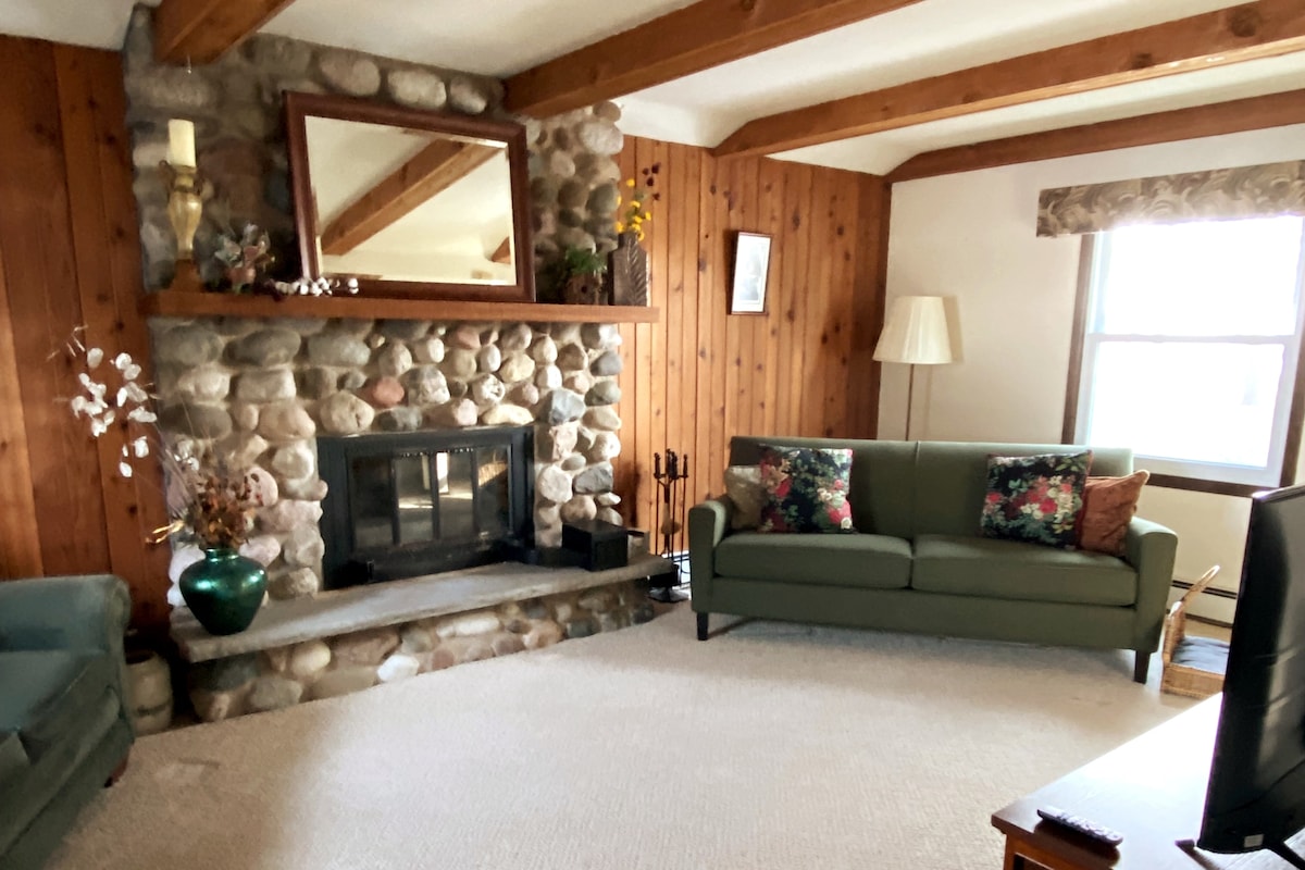 A cozy living area is equipped with two green sofas, each adorned with floral cushions. A stone fireplace is the focal point, complemented by a wooden mantel and a large mirror above it. Natural light filters in through a window dressed with a patterned valance.