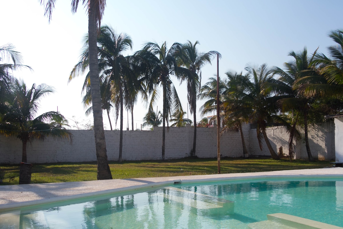 A clear view of a swimming pool is presented, surrounded by lush palm trees. A spacious grassy area lies beyond the pool, providing an open and natural setting. The sun shines brightly, casting gentle shadows across the scene.
