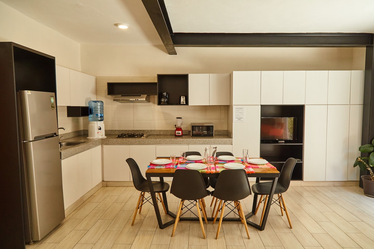 A modern kitchen area features a sleek design with light cabinetry and a large dining table set for six. The stainless steel refrigerator is positioned next to a water cooler. Appliances include a microwave, blender, and stove, enhancing functionality within the space.