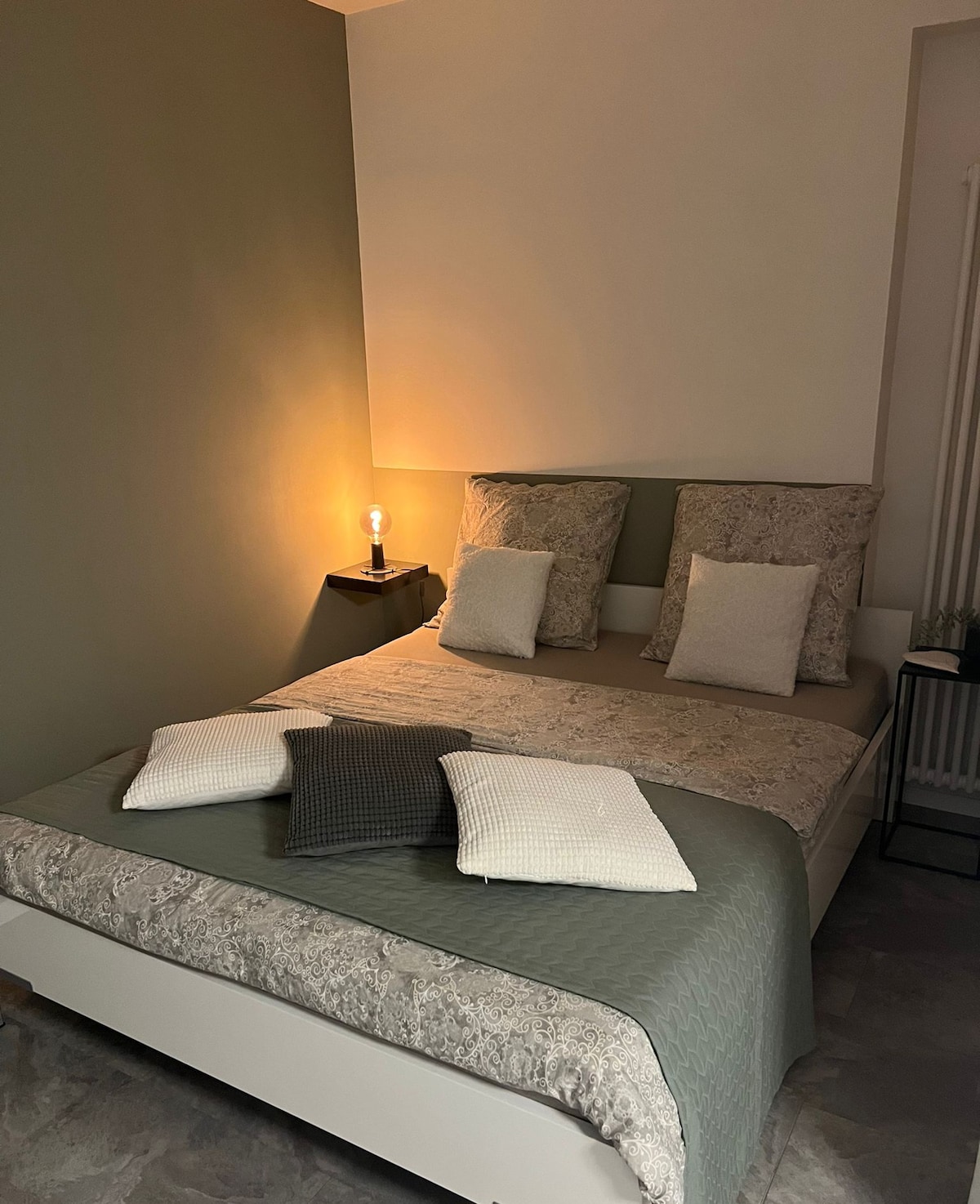 A cozy sleeping area features a spacious bed adorned with textured bed linens and decorative pillows in varying colors. Soft lighting from a bedside lamp creates a calm atmosphere, while the minimalist decor is complemented by a subtle green accent wall.
