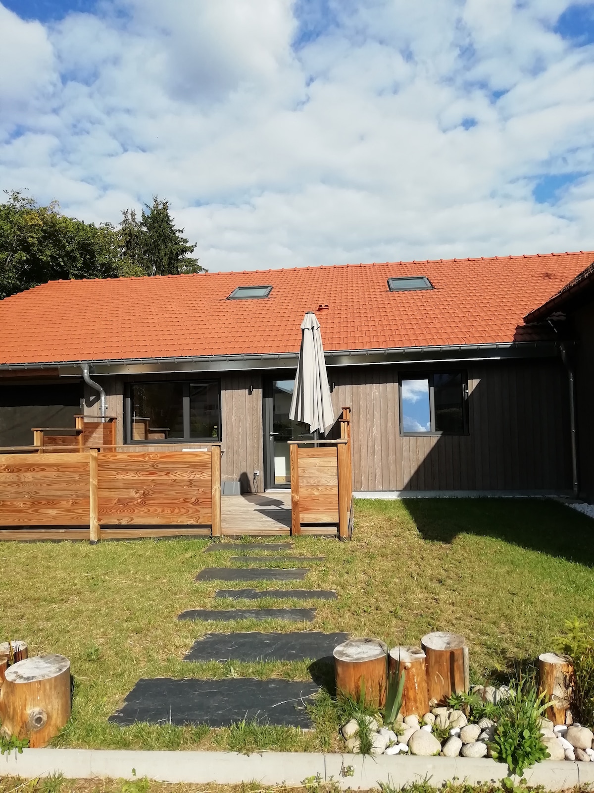 An exterior view of the gîte is presented, showcasing a wooden façade with a red roof. A pathway made of stone leads to the entrance, surrounded by a lawn and wooden logs. A patio with a central umbrella offers a relaxed outdoor space.