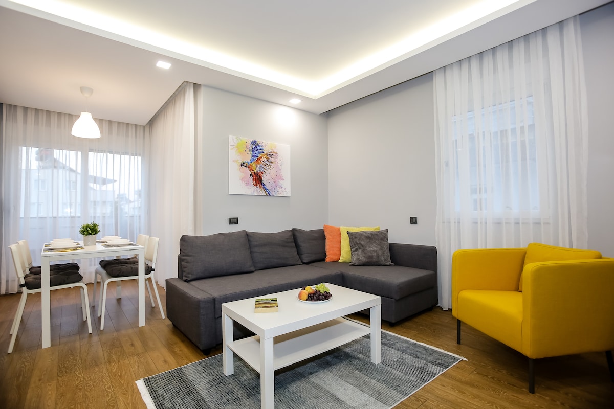 A modern living area is presented with a large gray sectional sofa featuring colorful cushions. A white coffee table sits at the center, adorned with a small fruit bowl. A dining table for four is visible in the background, alongside large windows allowing natural light to fill the room.