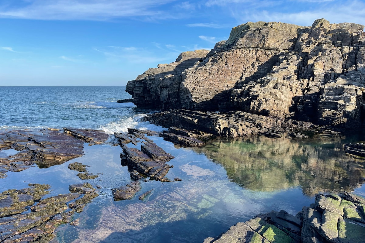 A rocky coastline is visible, showcasing layers of dark and light stones jutting out into the water. The calm sea reflects the clear blue sky, with gentle waves lapping against the rocks. The landscape conveys a natural and serene coastal environment.