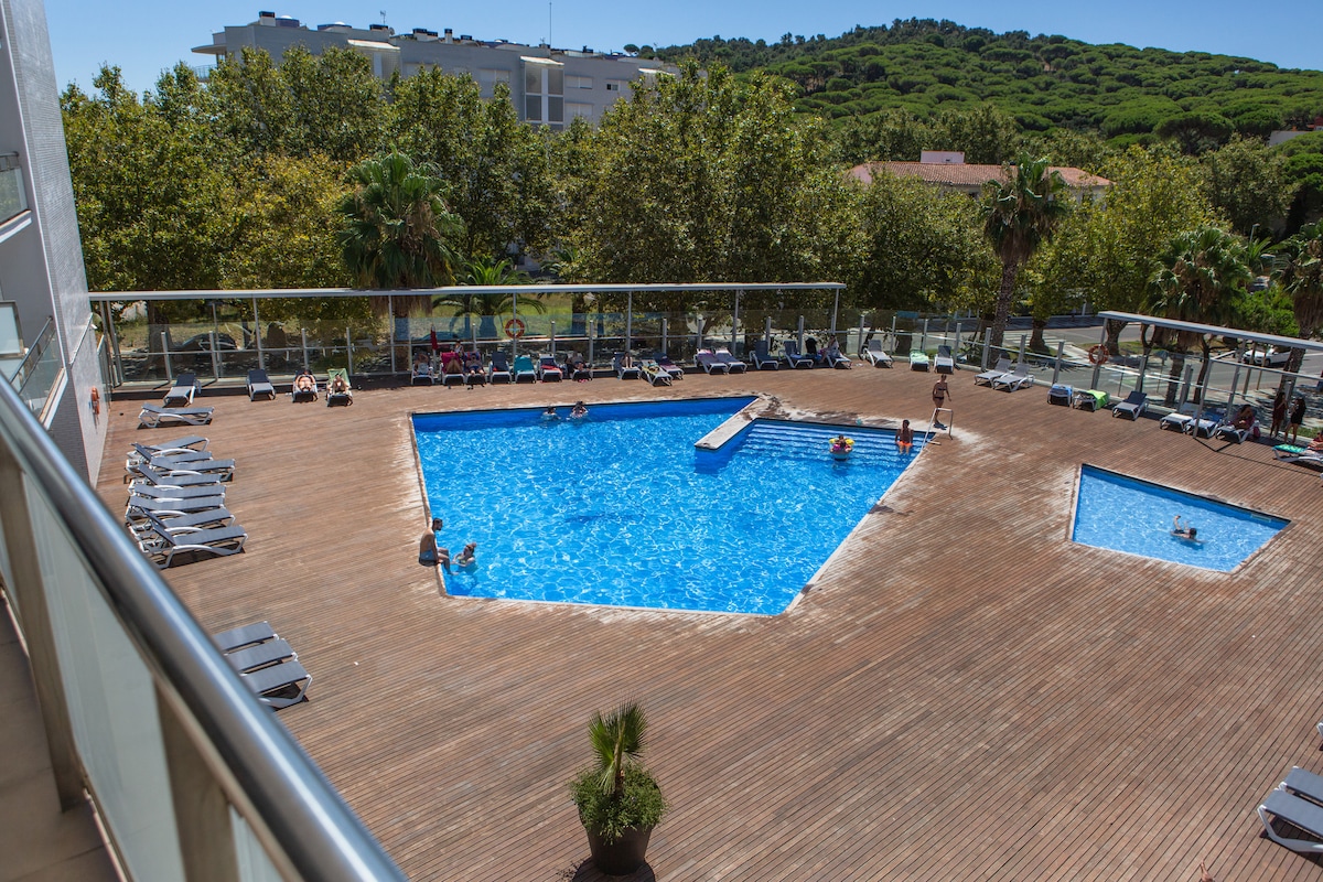 An expansive outdoor swimming pool is surrounded by a wooden deck, featuring varying depths and swim areas. Lounge chairs are arranged around the pool, with verdant trees providing shade. Sunlight reflects off the water, creating a bright and inviting atmosphere for relaxation.