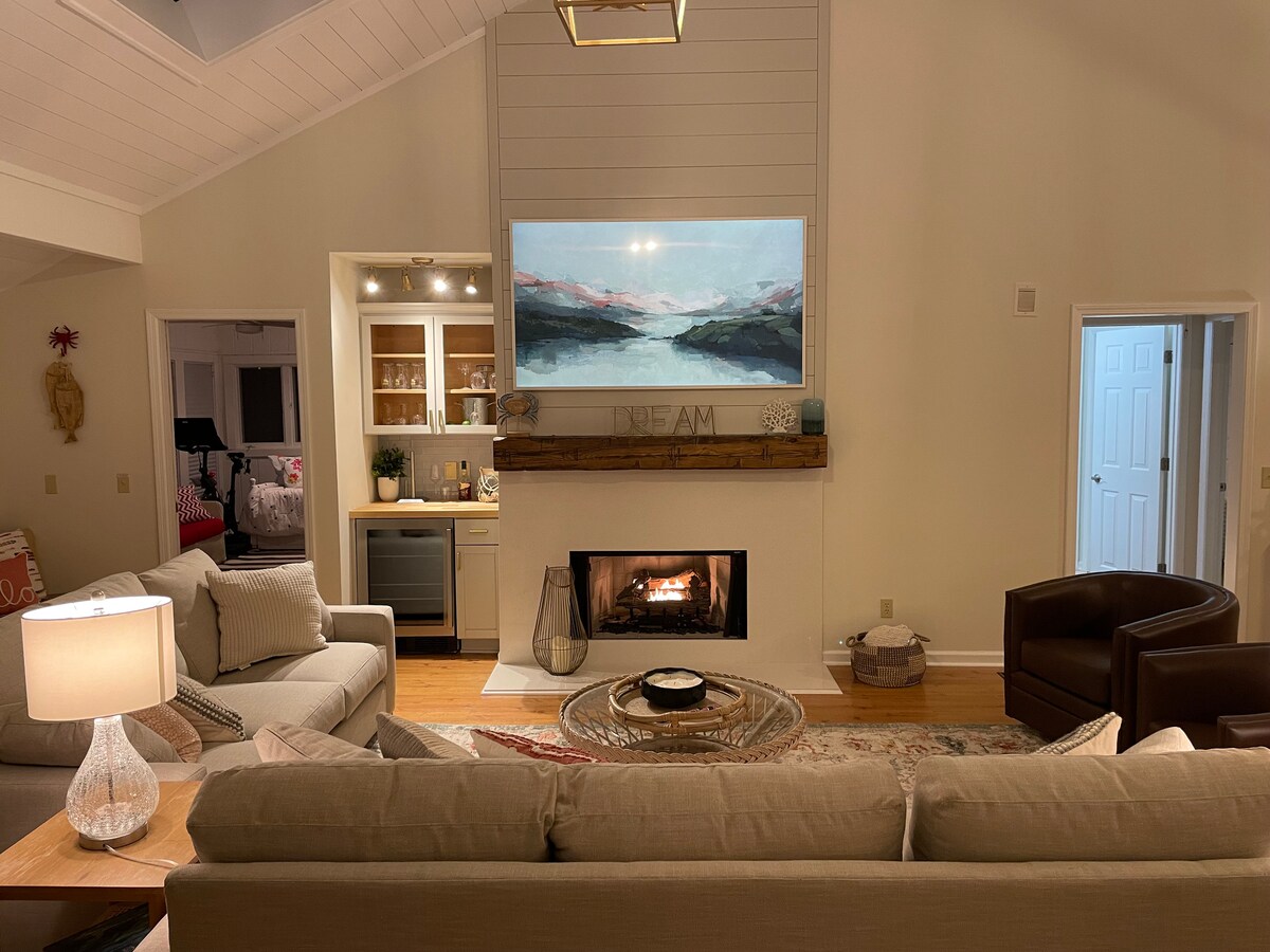 A cozy living area features a modern fireplace inset into a wall, with a large artwork depicting a serene landscape above it. Comfortable sofas and chairs are arranged around a central coffee table, offering a welcoming space for relaxation.