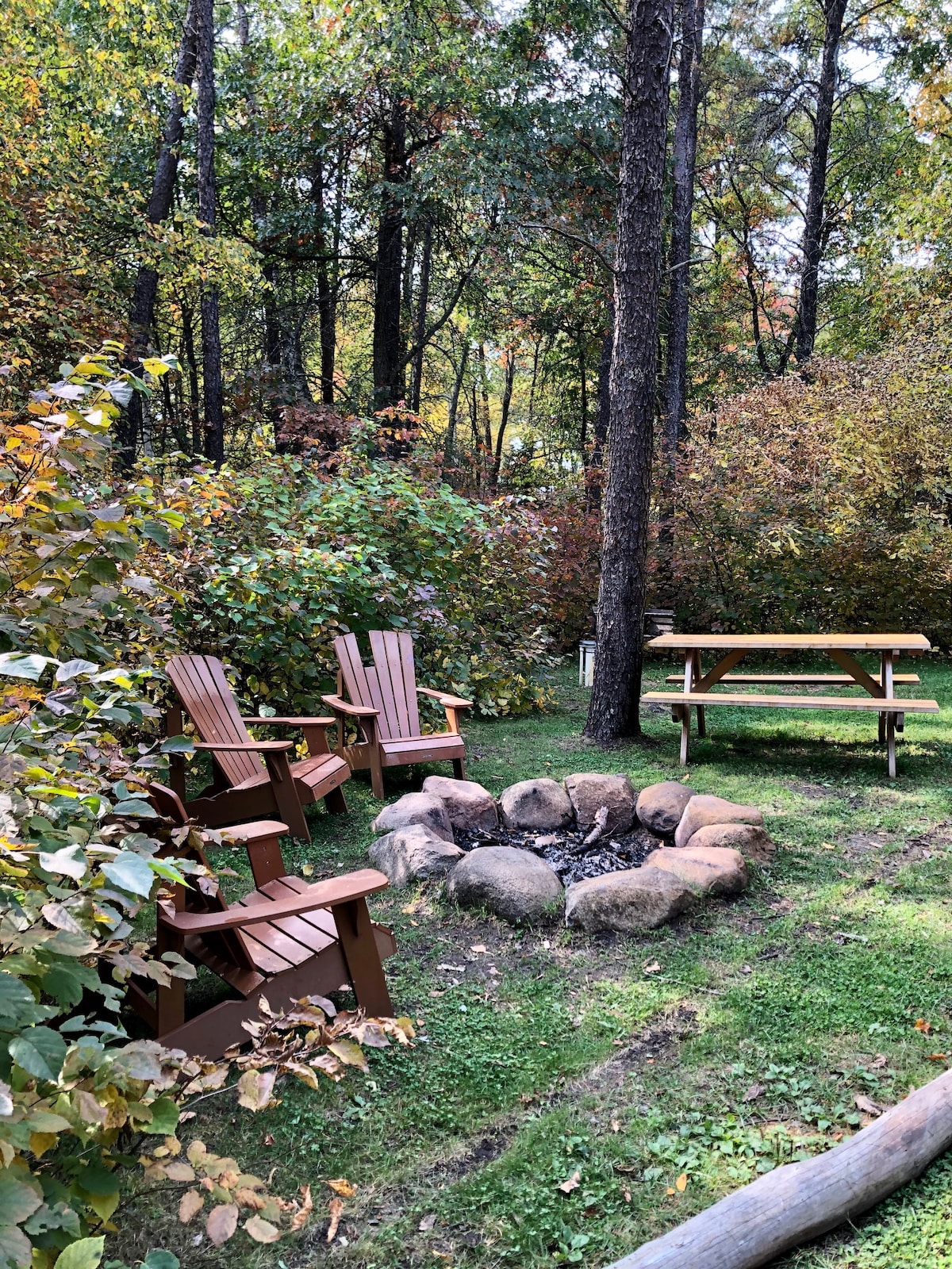 A fire pit area is surrounded by lush greenery and autumn foliage. Four wooden adirondack chairs are positioned around a circular arrangement of stones, while a picnic table sits nearby on a grassy patch, providing a cozy outdoor space for gatherings.