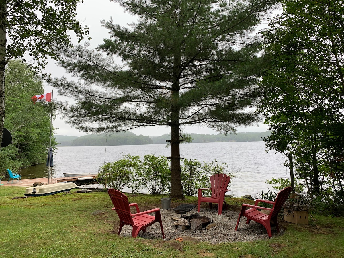A scenic riverside area features three red Adirondack chairs arranged around a fire pit, with a tall pine tree casting shade. A small dock extends into the lake, bordered by lush greenery and a Canadian flag gently waving in the breeze.