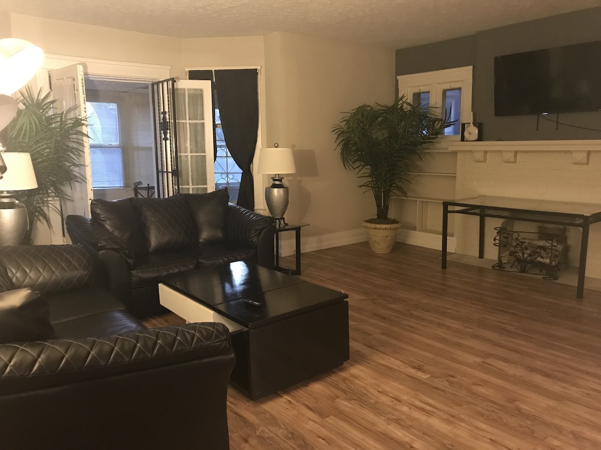 The living area features dark leather seating arranged around a coffee table. A wall-mounted television is visible, complemented by plants in decorative pots. Natural light enters through double doors, accentuated by contrasting dark curtains. The room maintains a clean and spacious appearance with wooden flooring.