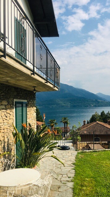 A stone exterior leads to a balcony with a metal railing, overlooking the serene landscape of Lake Como. A small table and chairs are situated on the stone pathway, surrounded by lush greenery, with mountains framing the background.