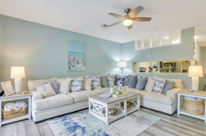 A spacious living area is furnished with a large sectional sofa adorned with patterned pillows. A light coffee table sits at the center, complemented by decorative elements. Soft lighting from lamps creates a cozy ambiance, while a view of the kitchen area can be seen in the background.