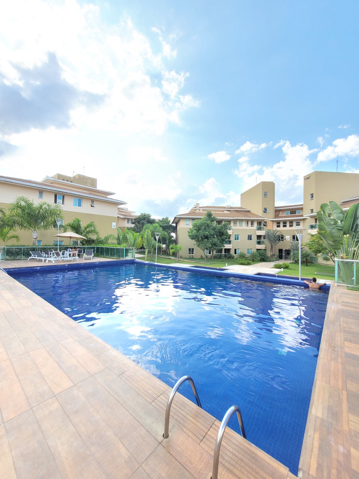 A well-maintained outdoor pool is surrounded by greenery and residential buildings. Clear blue water reflects the sky, and wooden decking provides a space for relaxation. Lounge chairs can be seen along the poolside, inviting guests to enjoy their time outdoors.