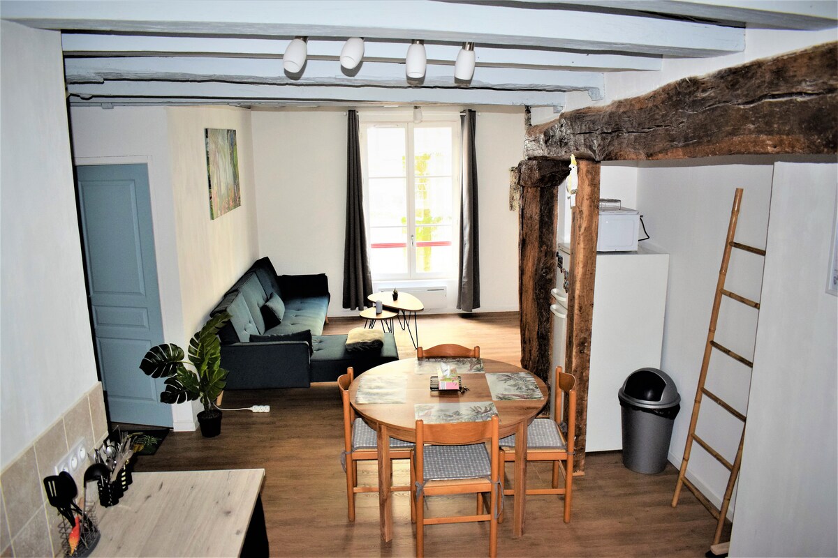 The open living area features a cozy seating arrangement with a dark sofa and a round dining table surrounded by six wooden chairs. Exposed wooden beams add character to the space, while natural light streams through the window with a distant view of the terrace.