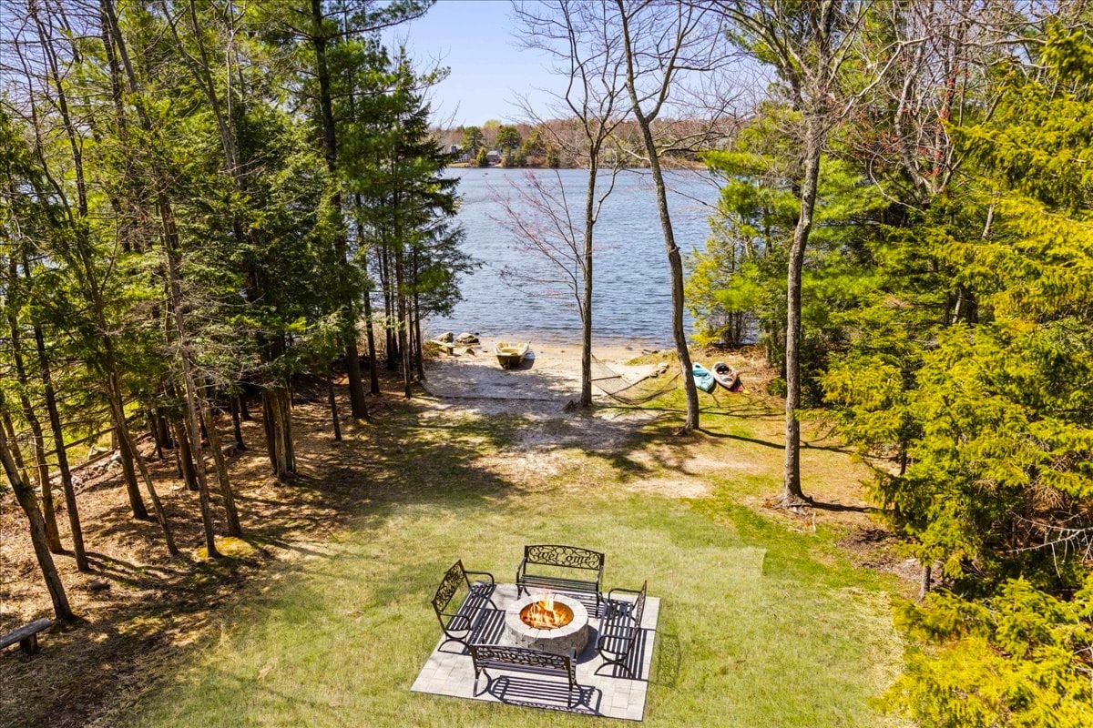 An inviting outdoor space features a fire pit surrounded by patio seating, set on a grassy area. Tall trees border the area, providing natural shade. The lake is visible in the background, with a sandy shoreline and kayaks nearby, suggesting opportunities for relaxation and recreation.