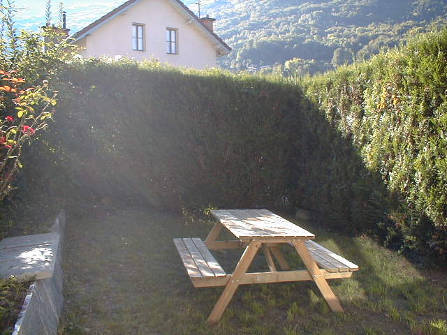 A wooden picnic table is set on a grassy area, surrounded by tall hedges. Sunlight filters through the foliage, creating a serene outdoor space. The backdrop features a gentle slope leading to a hillside, enhancing the tranquility of the scene.