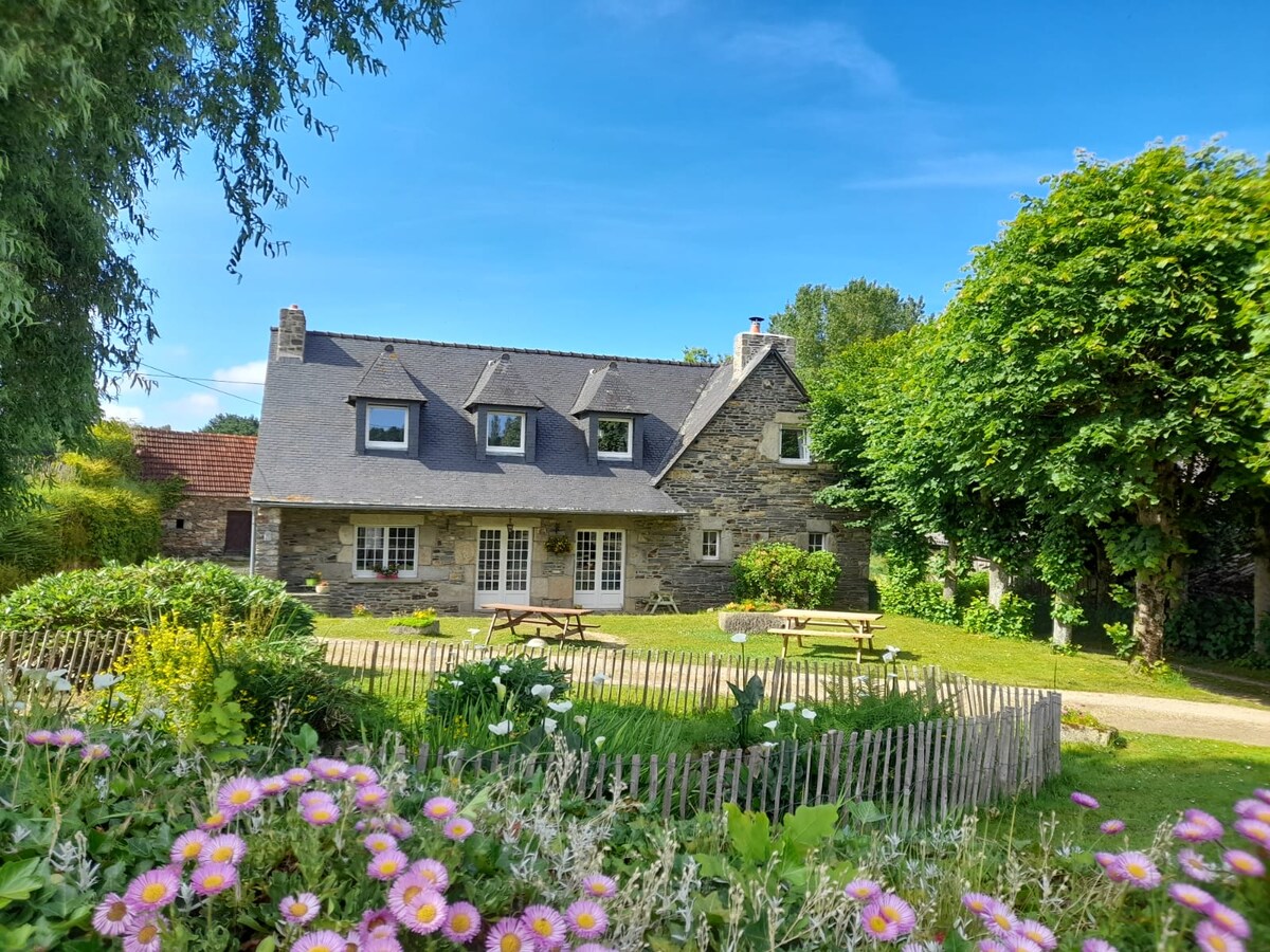 The exterior of a spacious countryside gîte is showcased, featuring stone walls and a slate roof. A well-maintained garden with colorful flowers is in the foreground. Several picnic tables are visible on the lush green lawn, surrounded by tall trees offering shade.