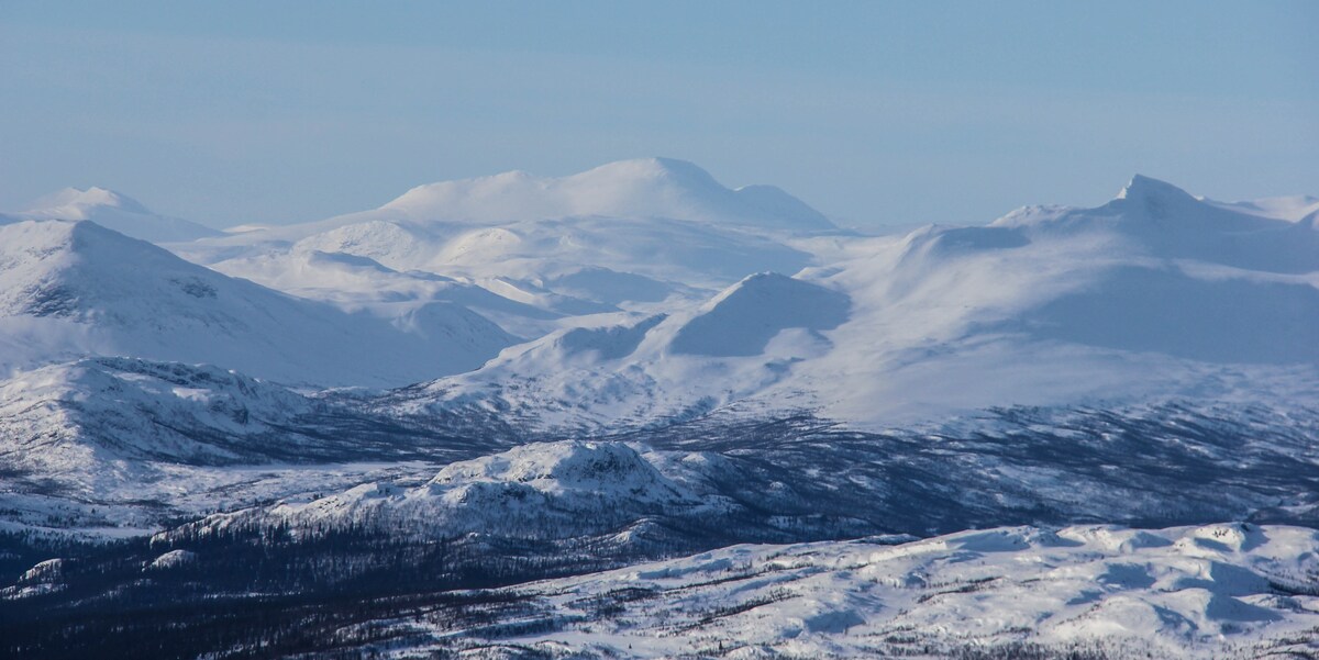 A panoramic view of snow-covered mountains stretches across the horizon, creating a serene winter landscape. The snow blankets the varied terrain, highlighting the contours of the landscape under a clear blue sky.