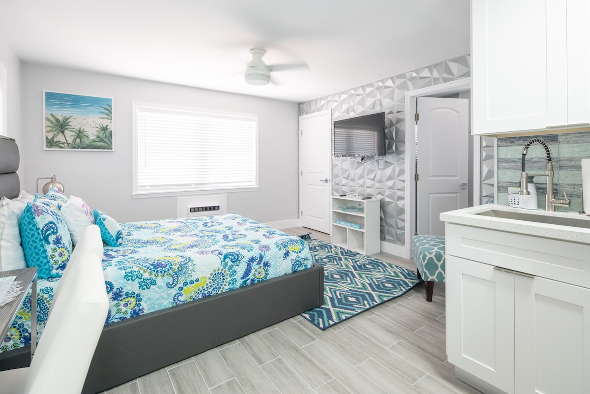 A modern studio is presented with a spacious bed covered in a colorful bedding set. A small kitchenette is visible, featuring sleek cabinetry and a sink. A patterned rug adds texture, while a television is mounted on the wall opposite the bed.