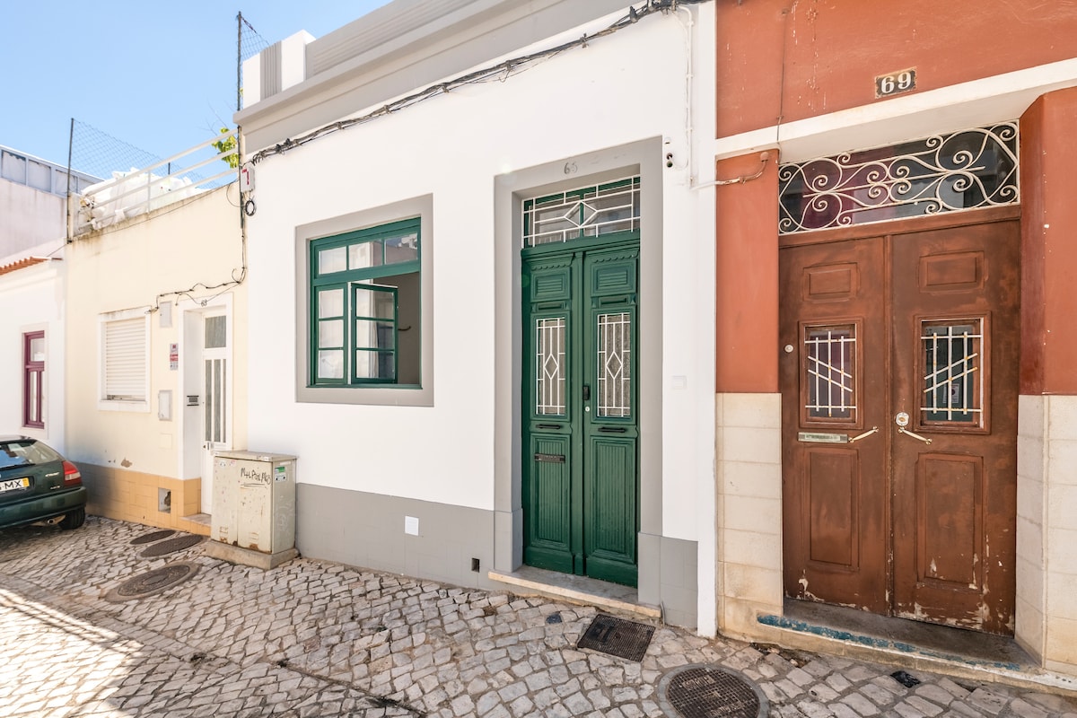 A traditional terraced house is showcased with a green double door and a window featuring white bars. The exterior is painted in soft hues, with cobblestone paving extending across the street. Nearby, an adjacent house exhibits a contrasting brown door, enhancing the charm of the neighborhood.