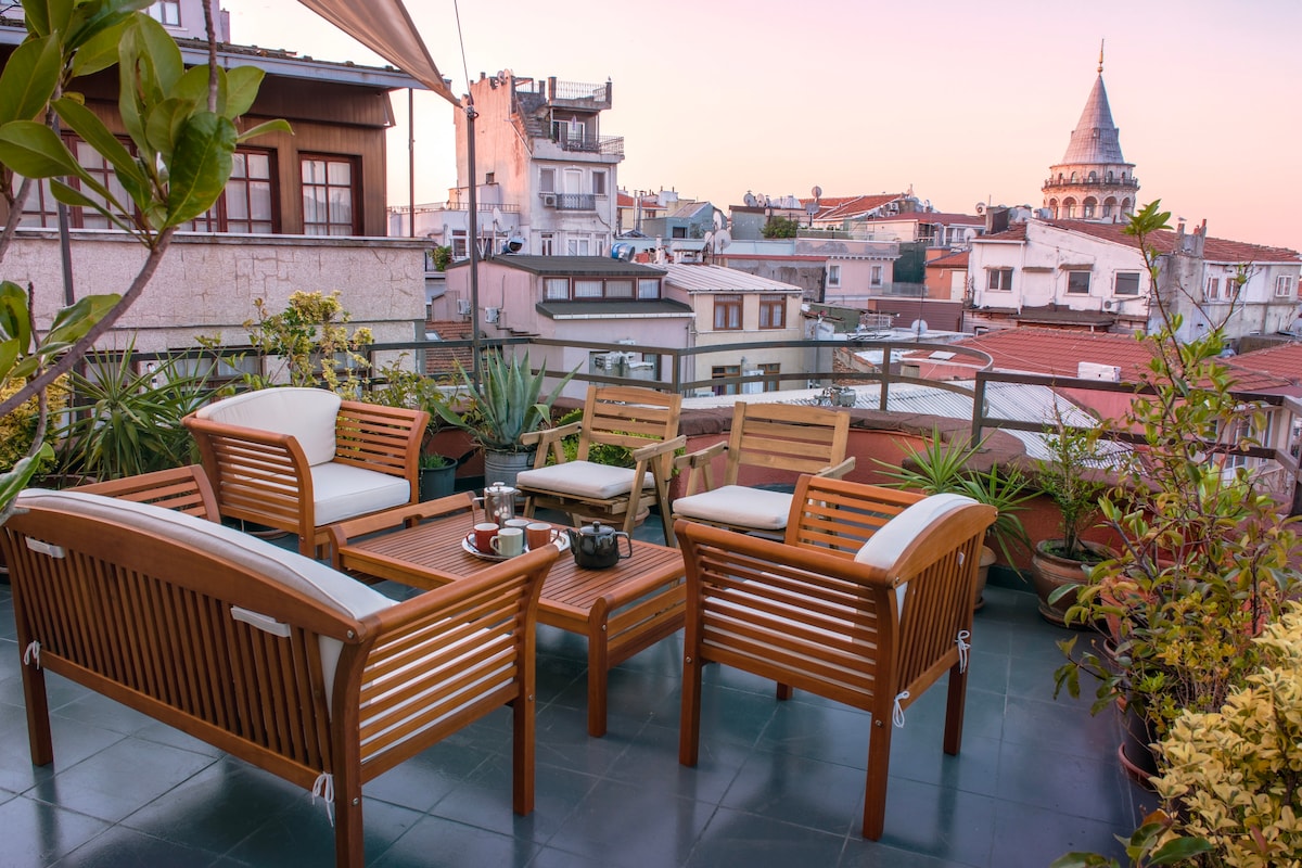A spacious private rooftop terrace features wooden seating and a coffee table, surrounded by vibrant plants. The view includes the rooftops of the city, with the iconic Galata Tower visible in the background against a colorful sunset sky.