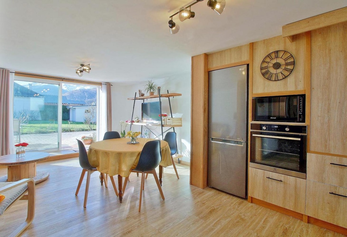 The open-concept living space features a dining table surrounded by four chairs. A modern kitchen area includes stainless steel appliances, complemented by wooden cabinetry. Large glass doors provide natural light and offer views of the garden and mountains beyond, creating an inviting area for relaxation.