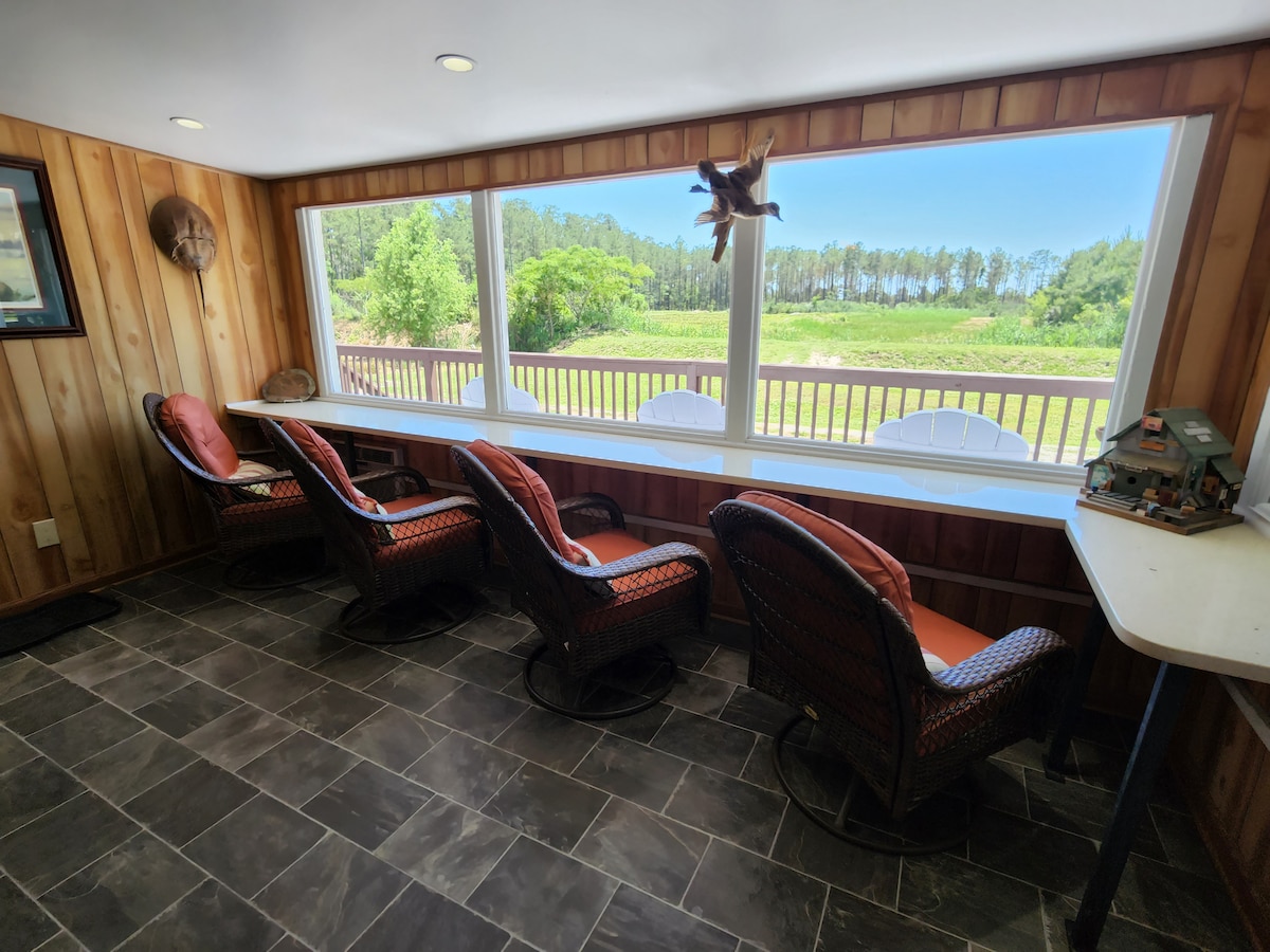 A sunlit all-season room features a long counter with seating, offering expansive views through large windows. Comfortable chairs are arranged around a table, while natural light highlights the rustic wooden walls and stone tile flooring.