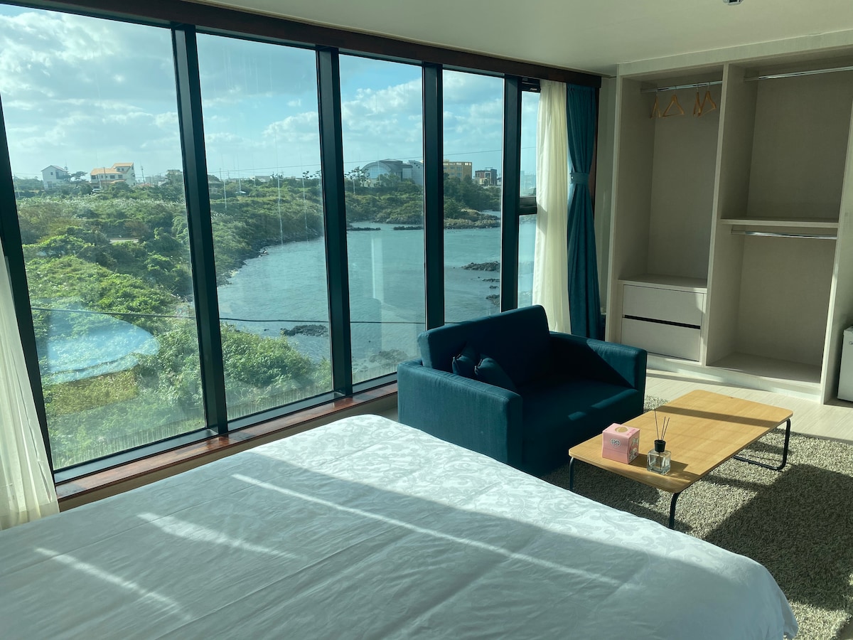 A spacious room is illuminated by natural light streaming through large floor-to-ceiling windows, showcasing a serene view of the water and greenery. A cozy seating area includes a blue couch and a small wooden coffee table, complementing the light-colored bedding in the foreground.