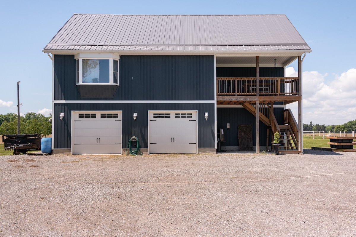 A two-story building is shown, featuring a sleek dark exterior and a covered balcony. Two garage doors are visible at ground level, flanked by outdoor lighting. Gravel leads to the entrance, with open fields beyond, highlighting the property’s countryside setting.