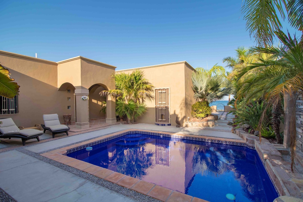 A serene outdoor space features a clear, unheated pool surrounded by tropical landscaping. Lounge chairs are positioned by the poolside, and a pathway leads to the entrance of the home. The water reflects the tranquil surroundings, enhancing the peaceful ambiance.
