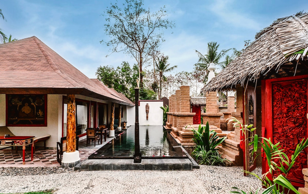 A serene outdoor area is captured, featuring a private plunge pool surrounded by lush greenery. Single-story structures with traditional design elements are present, including a thatched roof building and intricately carved walls. A natural stone pathway connects the spaces, enhancing the tranquil atmosphere.