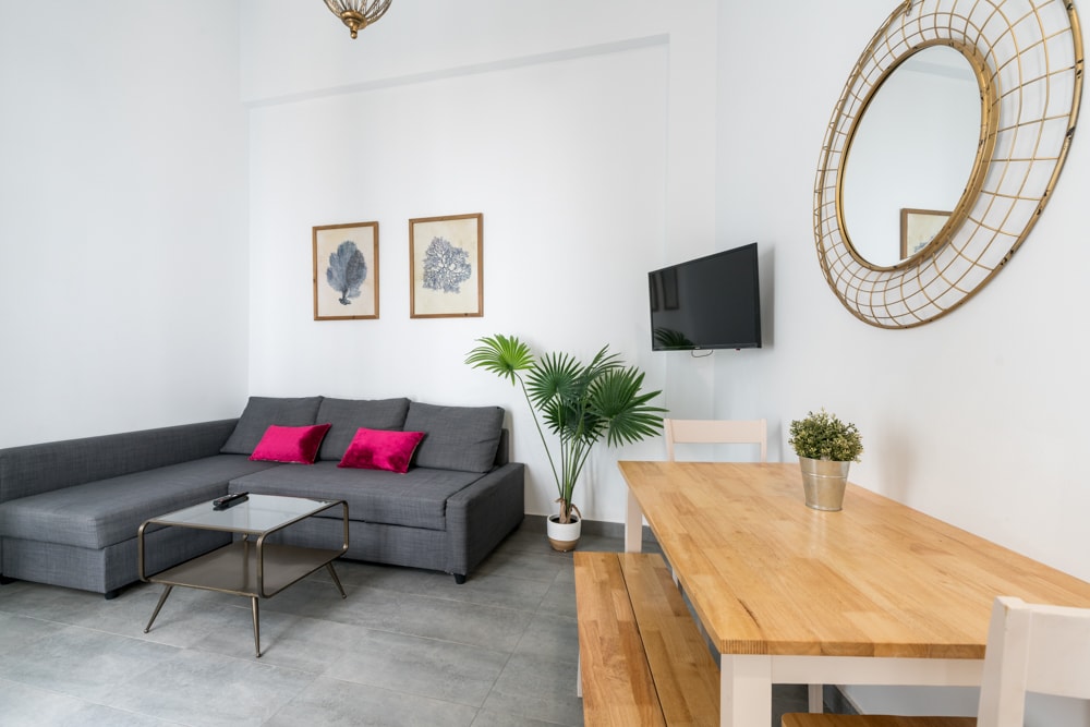 A bright living room features a gray sofa with pink cushions, a glass coffee table, and two framed botanical prints on the wall. A potted plant adds a touch of greenery, while a round, gold-framed mirror reflects natural light. A wooden dining table with white chairs is positioned nearby.