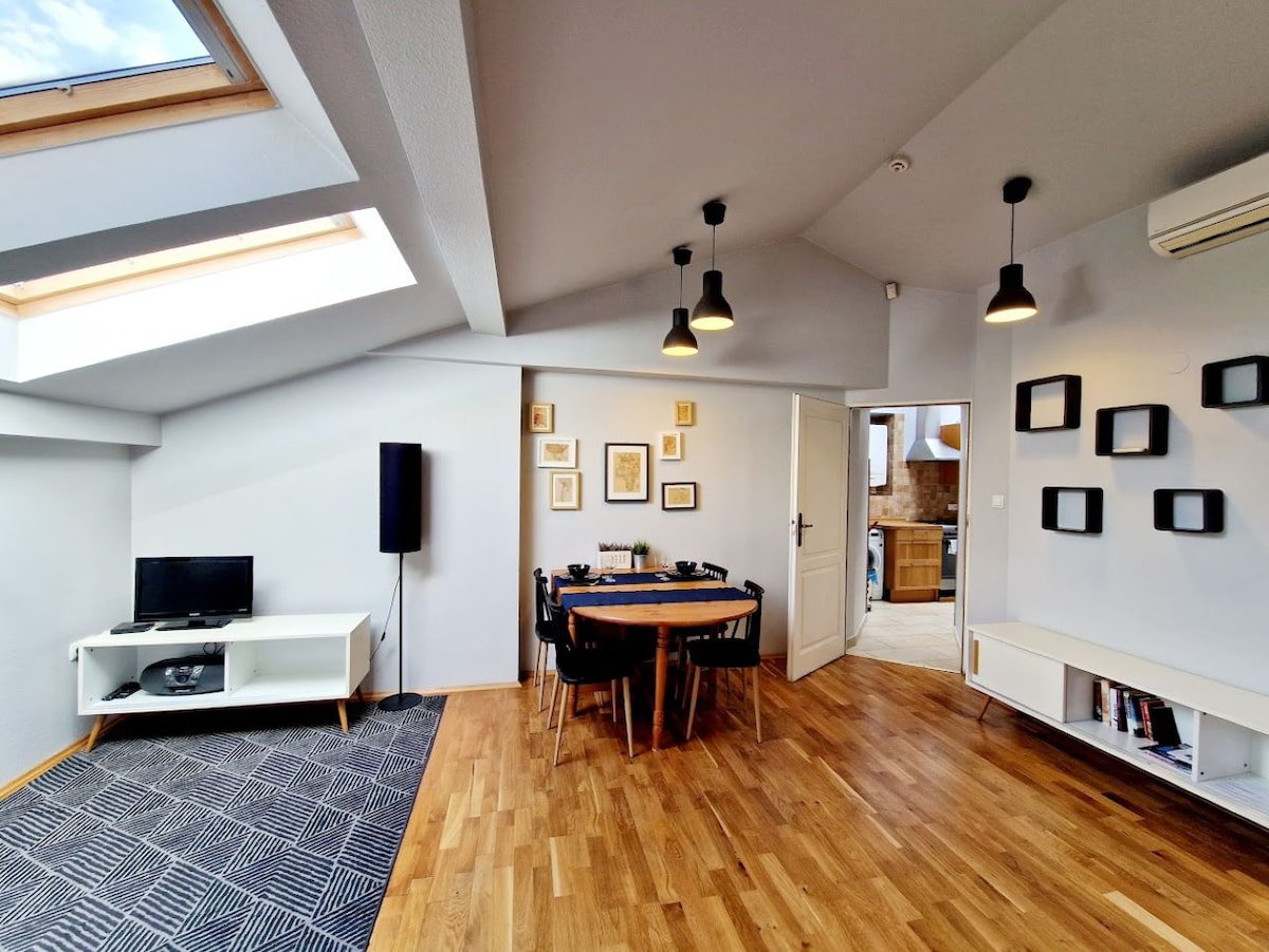 A modern living space features a round dining table surrounded by chairs, highlighted by natural light from skylights. A TV unit is positioned nearby, and a stylish black and white rug adds warmth to the wooden floor. Decorated walls showcase framed artwork.