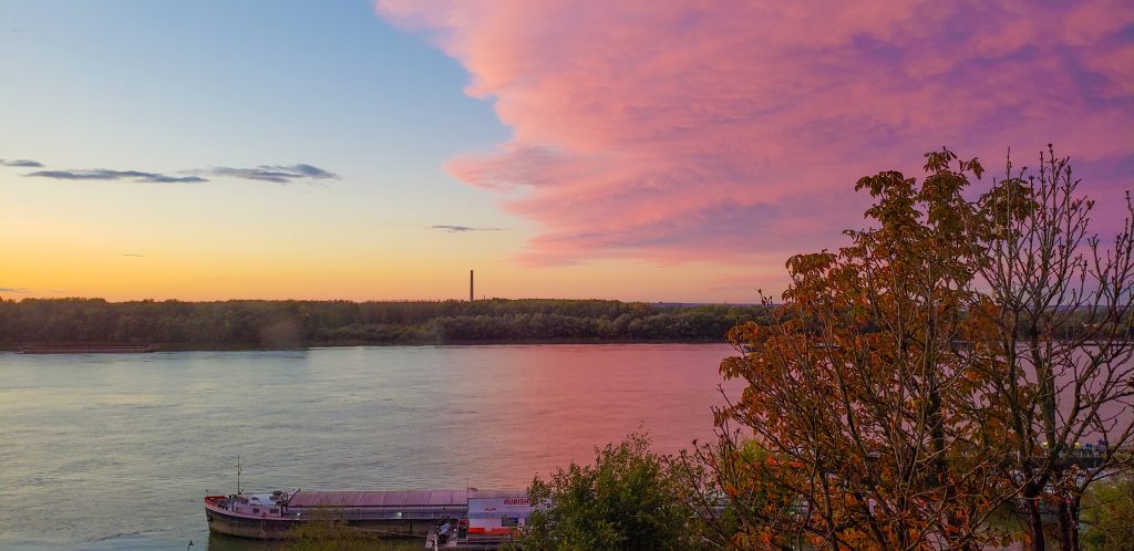 A scenic view of the Danube River at sunset, showcasing vibrant pink and orange clouds reflected on the water's surface. In the foreground, a tree with greenery is visible, alongside a moored boat along the riverbank. The distant horizon features lush trees.