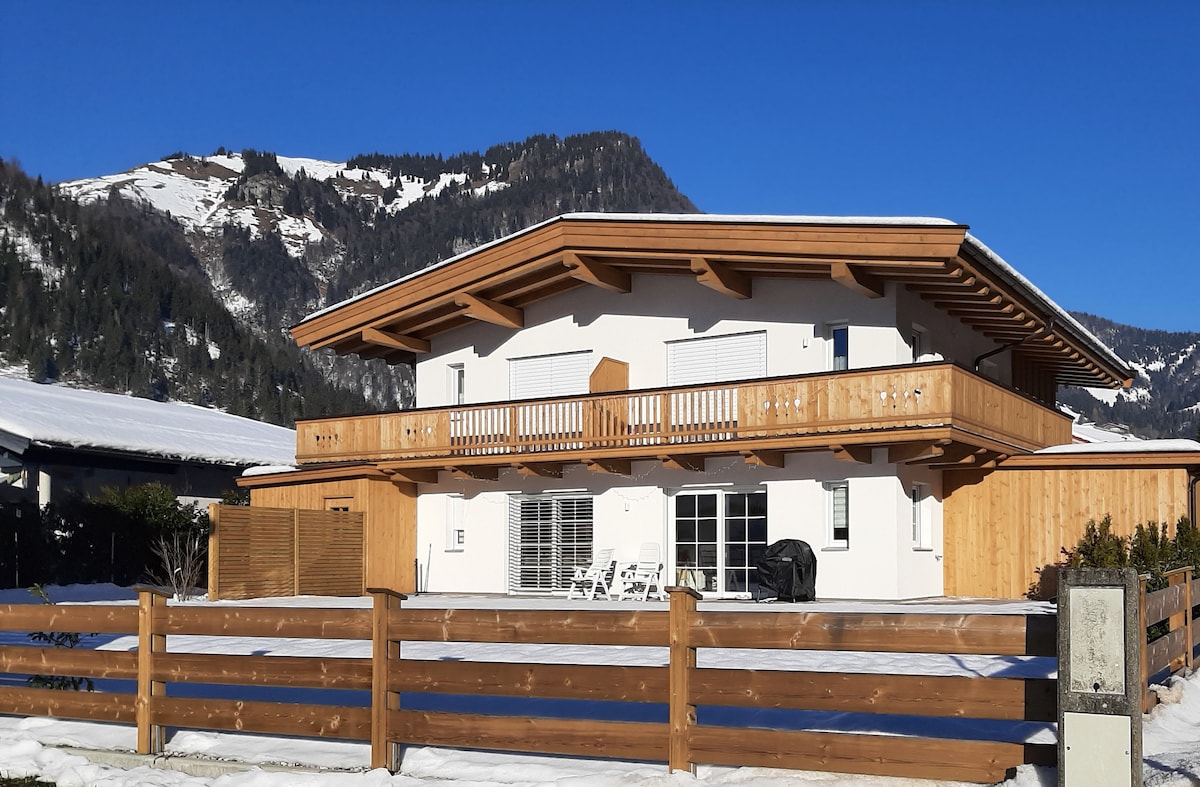 A modern two-story building with a wooden balcony overlooks a snowy landscape. Large windows reflect the sun's light, providing a view of the surrounding mountains. The exterior features wooden accents, with a fenced area and outdoor furniture visible.